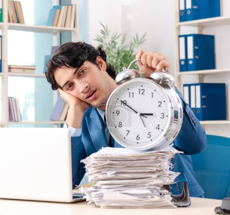 Foto de The young handsome male employee with too much work in the office - Imagen libre de derechos