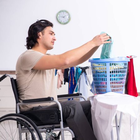 Téléchargez les photos : Le jeune homme en fauteuil roulant fait du repassage à la maison - en image libre de droit