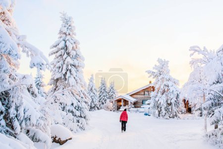 Foto de Vista trasera de una joven caminando entre chozas de madera y árboles cubiertos de nieve en Laponia Finlandia - Imagen libre de derechos