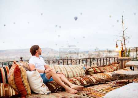 Téléchargez les photos : Jeune homme se relaxant sur la terrasse avec vue imprenable sur les montgolfières volant au-dessus de la ville de Goreme en Cappadoce Turquie - en image libre de droit