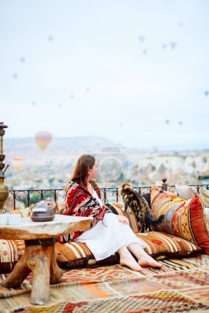 Téléchargez les photos : Young woman relaxing on terrace with amazing morning view of hot air balloons flying over Goreme town in Cappadocia Turkey - en image libre de droit