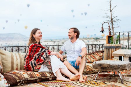 Foto de Hermosa pareja relajándose en la terraza con increíble vista de la mañana de globos de aire caliente volando sobre la ciudad de Goreme en Capadocia Turquía - Imagen libre de derechos