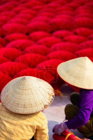 Téléchargez les photos : Deux travailleuses portant des chapeaux coniques parmi des bâtons d'encens rouges séchant à l'extérieur dans un village près de Hanoi au Vietnam - en image libre de droit