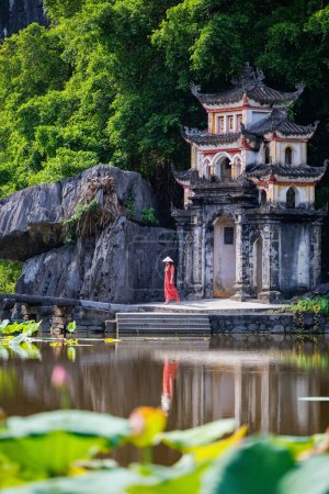 Téléchargez les photos : Belle femme en robe rouge et chapeau conique visitant la pagode Bich Dong à Ninh Binh Vietnam. Pagode Bich Dong est une destination touristique populaire de l'Asie. - en image libre de droit