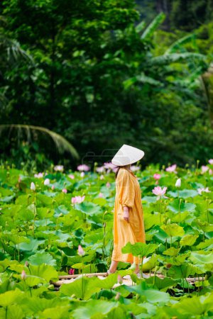 Foto de Hermosa mujer con vestido amarillo y sombrero cónico vietnamita rodeada de lago con flores de loto - Imagen libre de derechos