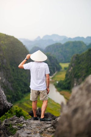 Foto de Vista posterior de un hombre joven con sombrero cónico disfruta de impresionantes vistas de las montañas kársticas y el río de la Cueva de Mua en Vietnam. - Imagen libre de derechos