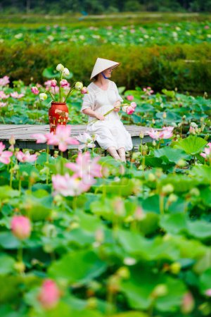 Téléchargez les photos : Belle femme portant une robe blanche et un chapeau vietnamien conique entouré d'un lac avec des fleurs de lotus - en image libre de droit