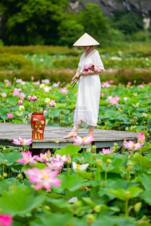 Foto de Hermosa mujer con vestido blanco y sombrero cónico vietnamita rodeada de lago con flores de loto - Imagen libre de derechos