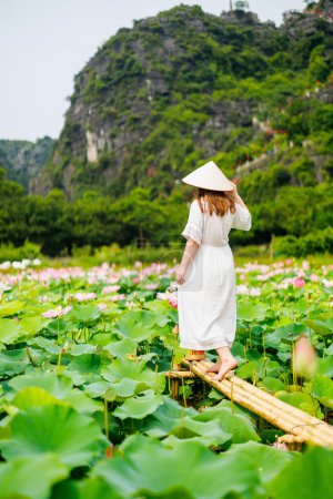 Foto de Hermosa mujer con vestido blanco y sombrero cónico vietnamita rodeada de lago con flores de loto - Imagen libre de derechos