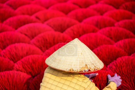 Téléchargez les photos : Travailleuse portant un chapeau conique parmi les bâtons d'encens rouges séchant à l'extérieur dans un village près de Hanoi au Vietnam - en image libre de droit