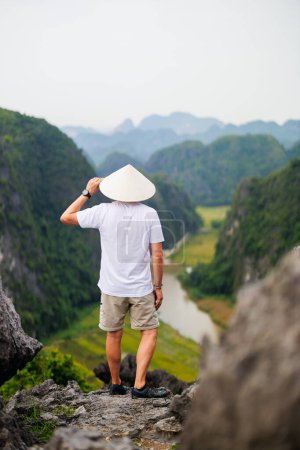 Back view of young man wearing conical hat enjoys stunning views of karst mountains and river of Mua Cave in Vietnam.