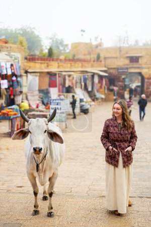 Téléchargez les photos : Femme caucasienne marchant à côté d'une vache dans une rue de la ville de Jaisalmer en Inde - en image libre de droit