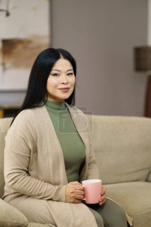 Niedliche asiatische MILF sitzt auf der Couch mit Tasse Kaffee nach Hause. Das Bild fängt Momente der Entspannung und Muße ein, während die Frau ihre Kaffeepause bequem von zu Hause aus genießt. . Hochwertiges Foto