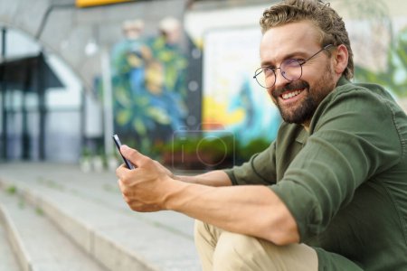 Man sitting on stone stairs on street, using pocket PC. With compact and portable device in hand, he effortlessly combines technology with surroundings. urban backdrop adds modern touch to scene. High
