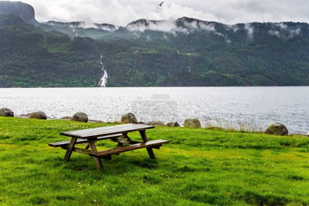 Foto de Mesa con bancos para picnic junto al gran lago junto a la carretera a Roldal. Pintoresco paisaje en el oeste de Noruega. Día de verano frío y lluvioso. Paisaje escénico en Noruega Occidental. - Imagen libre de derechos