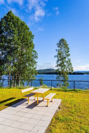 Mesa de picnic y bancos junto al lago. Acogedor rincón para turistas. Mañana mágica de verano. Nubes blancas claras en el cielo azul iluminadas por el amanecer. Cálido julio en Noruega. 