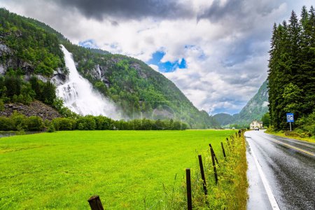 Photo for Roaring high-water cascading waterfall Vidfossen. Lush clouds and constant cold rain. Scenic road in the mountains of Western Norway. White water foam envelops the foot of the waterfal - Royalty Free Image
