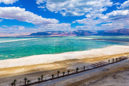  Dead Sea. Israel. The water reflects the bright blue of the sky. Lush cumulus clouds are reflected in the water. Drone footage. Haghway lined with palm trees runs along the seashore.