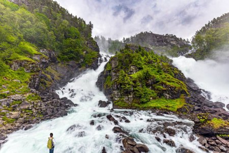 Woman admiring a picturesque double waterfall Lotefossen. Travel to NorwaySummer rainy day. The road passes through water jets. Cloud of water dust hangs in the air.