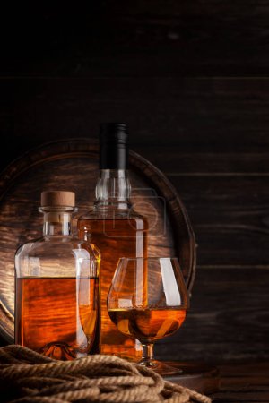 Téléchargez les photos : Verre et bouteilles de cognac, whisky ou rhum doré. Devant un vieux tonneau en bois avec espace de copie - en image libre de droit