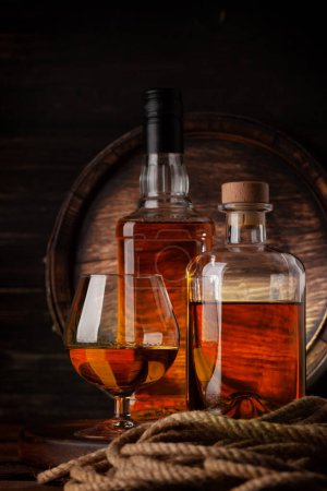 Photo for Glass and bottles with cognac, whiskey or golden rum. In front of old wooden barrel - Royalty Free Image