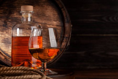 Photo for Glass and bottle with cognac, whiskey or golden rum. In front of old wooden barrel with copy space - Royalty Free Image