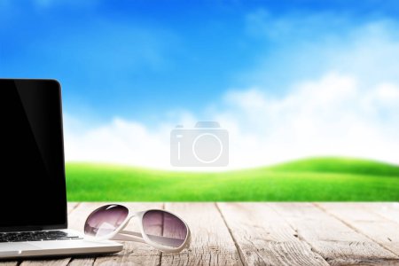 Téléchargez les photos : Ordinateur portable et lunettes de soleil sur une table en bois devant un paysage ensoleillé. Travail et voyages ou concept d'entreprise à distance - en image libre de droit