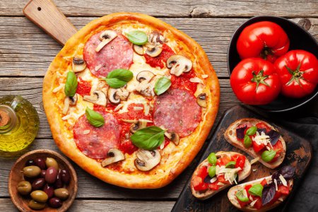 Foto de Cocina italiana. Pizza de pepperoni, aceitunas y tostadas antipasto. Colocación plana sobre mesa de madera - Imagen libre de derechos