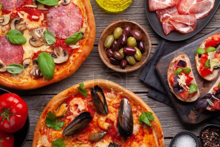 Foto de Cocina italiana. Pizza de pepperoni y mariscos, aceitunas y tostadas antipasto. Colocación plana sobre mesa de madera - Imagen libre de derechos