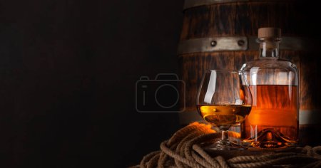 Téléchargez les photos : Verre et bouteille avec cognac, whisky ou rhum doré. Devant un vieux tonneau en bois avec espace de copie - en image libre de droit