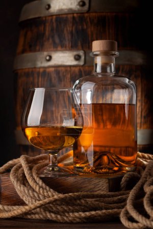 Téléchargez les photos : Verre et bouteille avec cognac, whisky ou rhum doré. Devant un vieux tonneau en bois - en image libre de droit