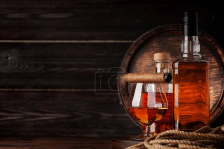 Téléchargez les photos : Verre et bouteille avec cognac, whisky ou rhum doré. Devant un vieux tonneau en bois avec espace de copie - en image libre de droit