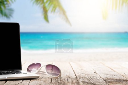 Photo for Laptop and sunglasses on wooden table in front of sunny sea and palm leaves. Work and travel or remote business concept - Royalty Free Image