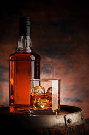 Photo for Glass of whiskey with ice cubes and bottle on the old barrel. With copy space on wooden background - Royalty Free Image