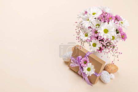 Foto de Caja de regalo, huevos de Pascua y ramo de flores sobre un fondo beige con espacio para sus saludos - Imagen libre de derechos