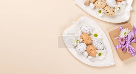 Téléchargez les photos : Oeufs de Pâques et fleurs sur assiette et boîte cadeau sur fond beige. Pose plate avec espace de copie - en image libre de droit