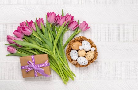 Foto de Pink tulip flowers bouquet, gift box and easter eggs. On white wooden table. Flat lay - Imagen libre de derechos