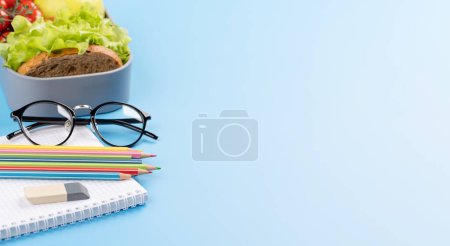 Foto de School supplies, stationery, and lunch box on blue background. Education and nutrition. With blank space for your text - Imagen libre de derechos