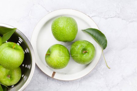 Photo for Fresh green apples in colander and on plate on stone table. Flat lay - Royalty Free Image
