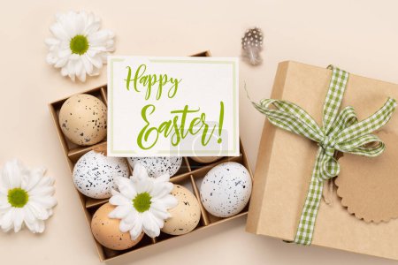 Foto de Caja de regalo, huevos de Pascua y flores sobre fondo beige con tarjeta de felicitación. Puesta plana - Imagen libre de derechos