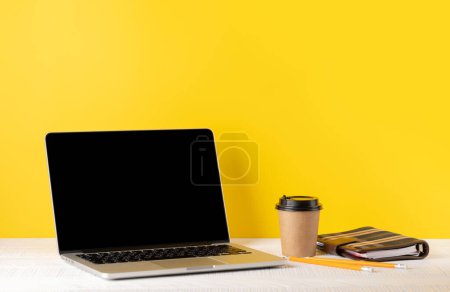 Photo for Laptop with blank screen for your message, app or web, coffee cup and notepad. Computer with black display. Over yellow background - Royalty Free Image