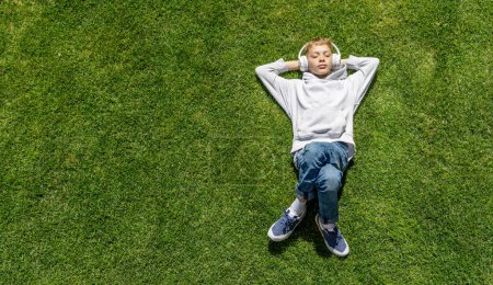 Téléchargez les photos : Un garçon se relaxant sur l'herbe, écoutant de la musique avec des écouteurs. Pose plate - en image libre de droit