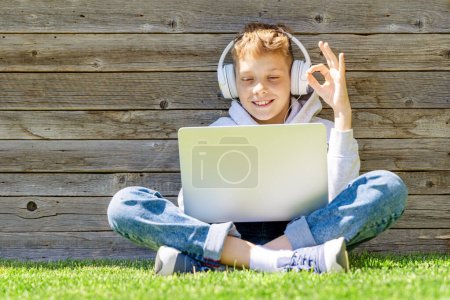 Téléchargez les photos : Un garçon utilisant un ordinateur portable et un casque à l'extérieur. Concept d'éducation ou de divertissement - en image libre de droit