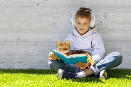 Téléchargez les photos : Un garçon avec un chat lisant un livre sur un champ herbeux - en image libre de droit