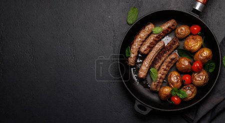 Téléchargez les photos : Délicieuses saucisses grillées et pommes de terre dans une poêle à frire pétillante. Pose plate avec espace de copie - en image libre de droit