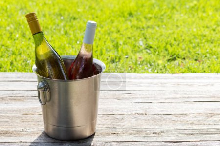Téléchargez les photos : Bouteilles de vin blanc et rose dans un seau à glace sur une table de jardin extérieure, préparant le terrain pour une délicieuse fête d'été. Avec espace de copie - en image libre de droit