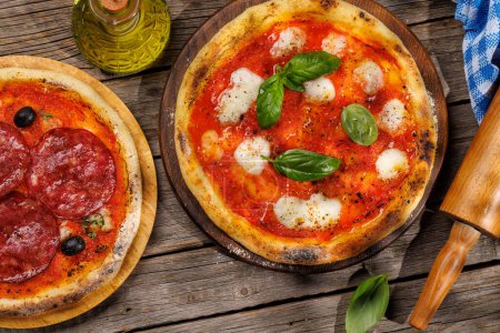 Foto de Pizza casera de margarita y pepperoni, cubierta con tomates frescos, queso mozzarella y hojas aromáticas de albahaca. En la mesa de jardín al aire libre plana - Imagen libre de derechos