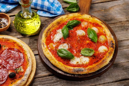 Foto de Pizza casera de margarita y pepperoni, cubierta con tomates frescos, queso mozzarella y hojas aromáticas de albahaca. En la mesa de jardín al aire libre - Imagen libre de derechos