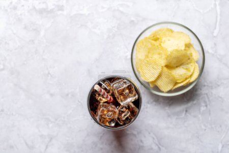 Photo for Refreshing glass of cola with ice, accompanied by a serving of crispy chips. On stone table with copy space. Flat lay - Royalty Free Image