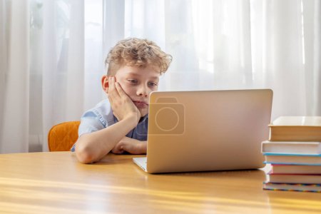 Photo for Thoughtful schoolboy studying online on laptop at desk with books and stationery - Royalty Free Image